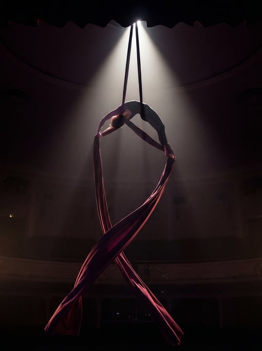 Aerial silk performer wrapped in burgundy fabric high above a dark stage