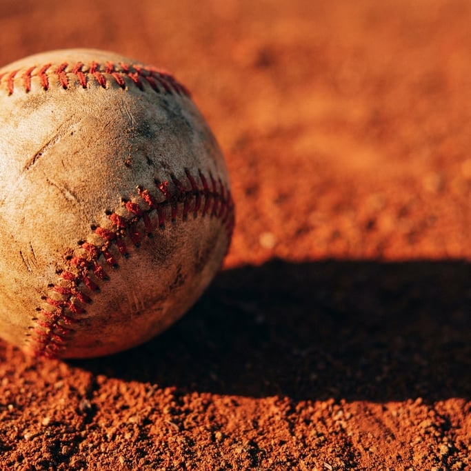Close-up of a worn baseball on red infield dirt, the stitching visible in sharp detail