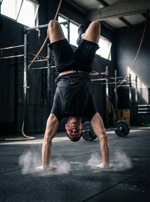 Handstand walk across a gym floor, athlete inverted, arms driving forward, shoulders loaded