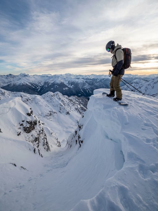 Snowboarder standing at the edge of a cornice looking down a steep chute, the drop visible below