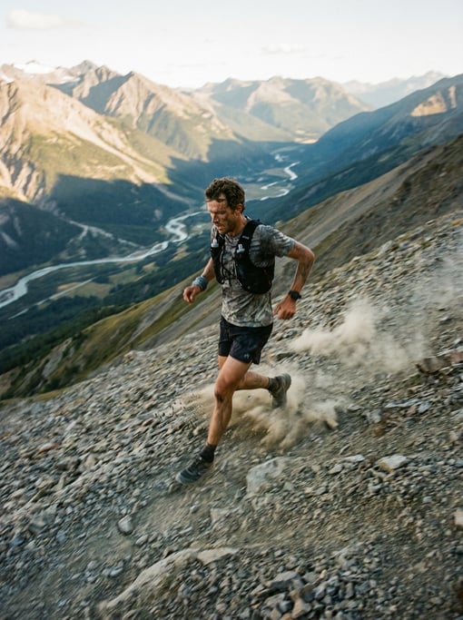 Trail runner descending a steep mountain trail with scree, dust rising from each foot strike