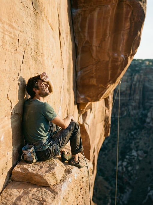 Rock climber resting on a ledge mid-route, looking up at the remaining climb, chalk bag on hip