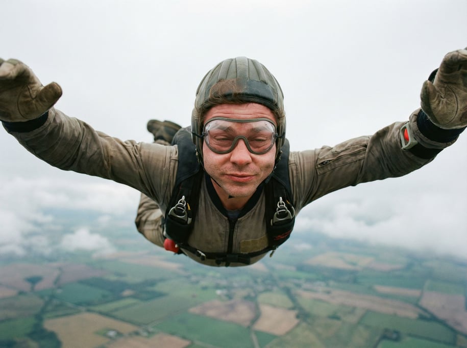 A skydiver in freefall, arms spread, earth a patchwork quilt far below, wind distorting cheeks (ly5tck)
