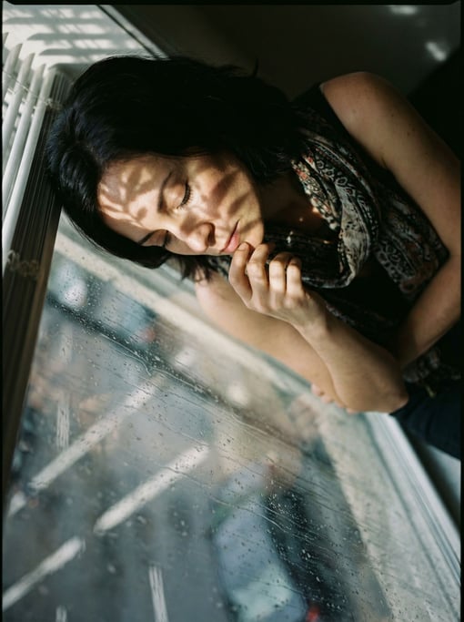 A woman in her 30s deep in thought at a window, face half-lit, rain streaking the glass beside her (nmslndh8)