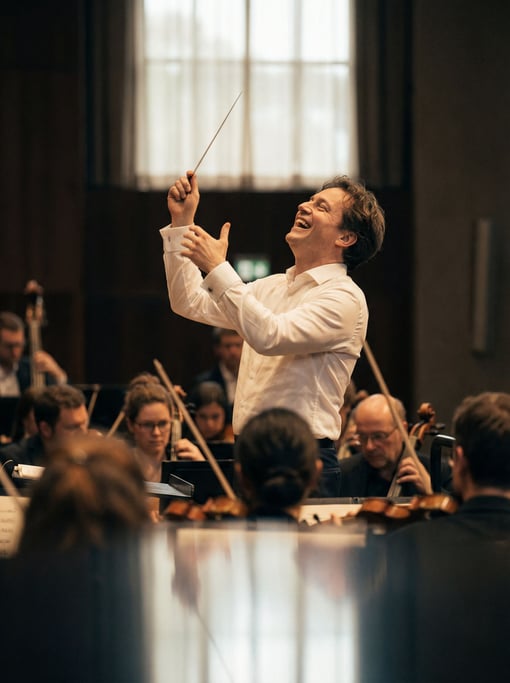 A conductor mid-gesture, baton frozen at the peak, orchestra blurred below, concert hall light