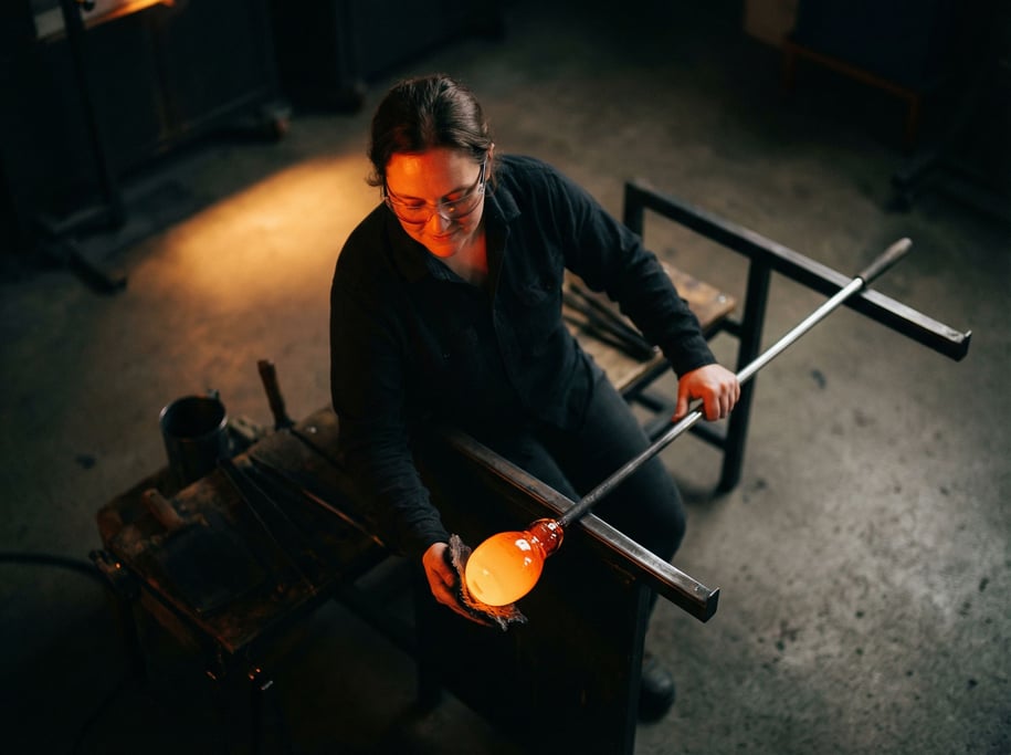A glassblower shaping molten glass on a punty rod, face lit by the orange glow of the gather (womznh)