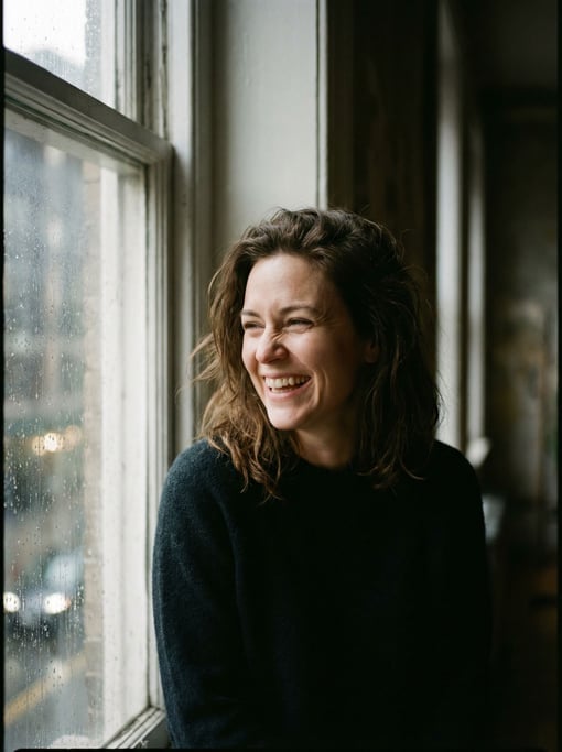 A woman in her 30s deep in thought at a window, face half-lit, rain streaking the glass beside her (afh17ezm)
