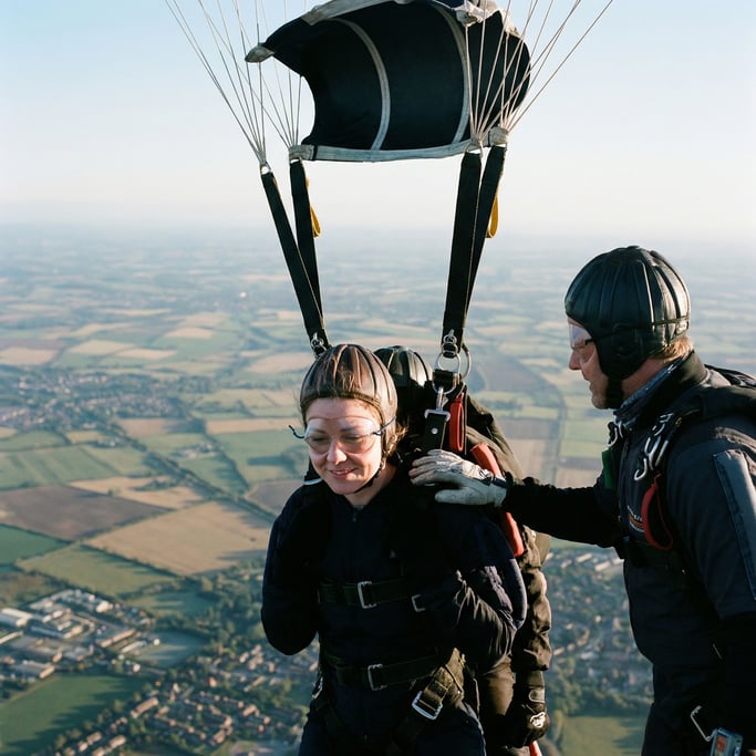 A skydiver in freefall, arms spread, earth a patchwork quilt far below, wind distorting cheeks (mi5q8tnu)