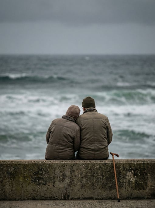An elderly couple sitting side by side on a seaside concrete wall, their backs to the camera