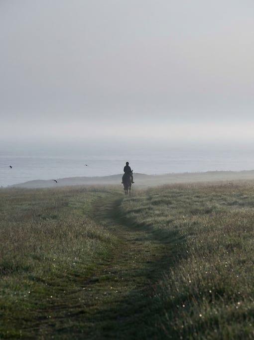 A woman in her 40s riding a horse alone through a misty coastal field at dawn