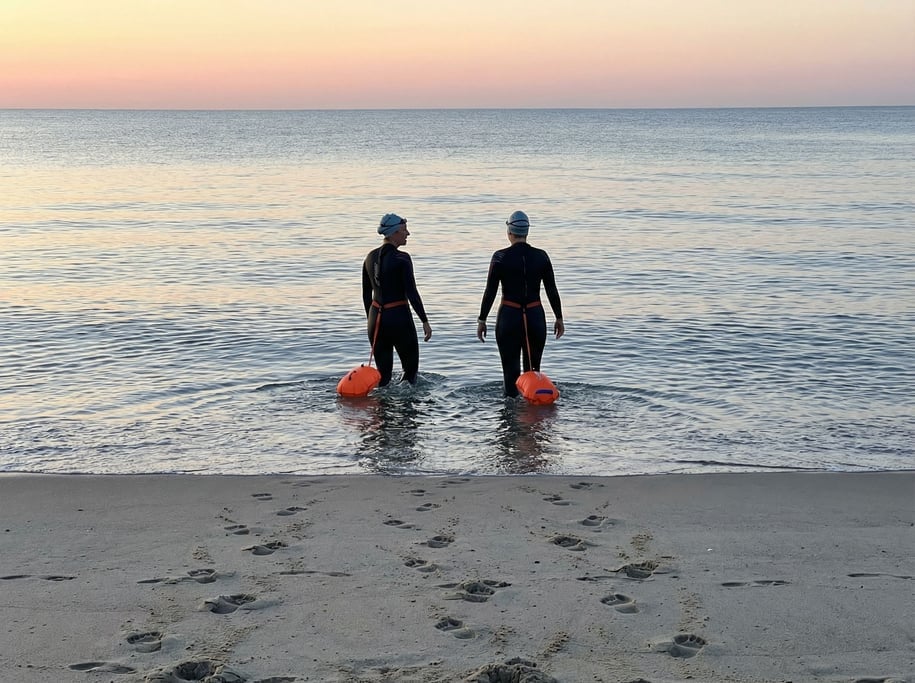 Two women in wetsuits walking into the ocean at dawn for an open-water swim