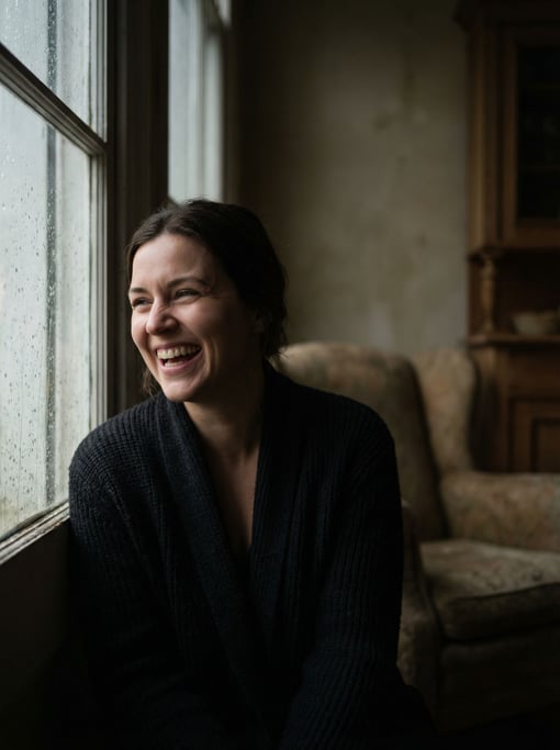A woman in her 30s deep in thought at a window, face half-lit, rain streaking the glass beside her