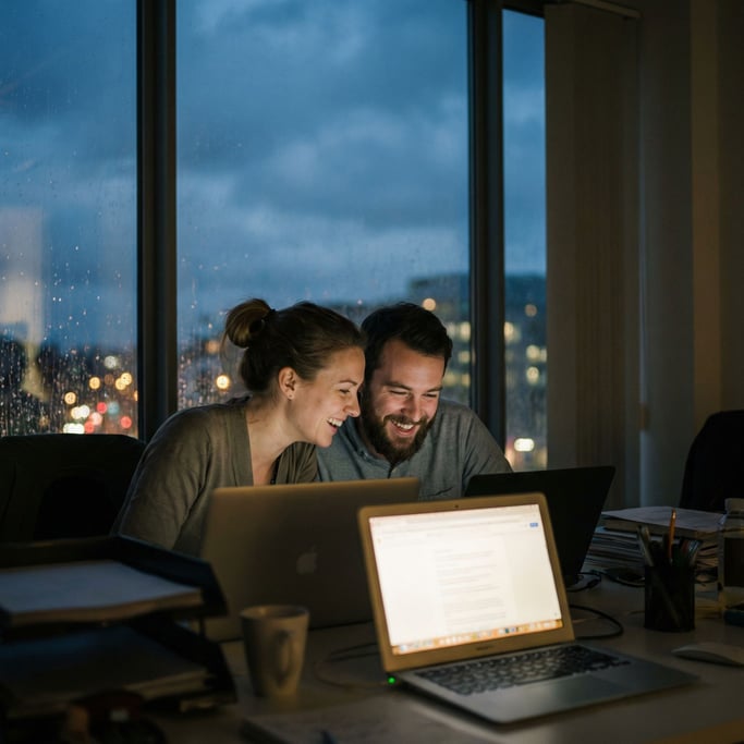 Two people working late, soft screen glow on their faces, city lights through the window behind (jvy4segc)