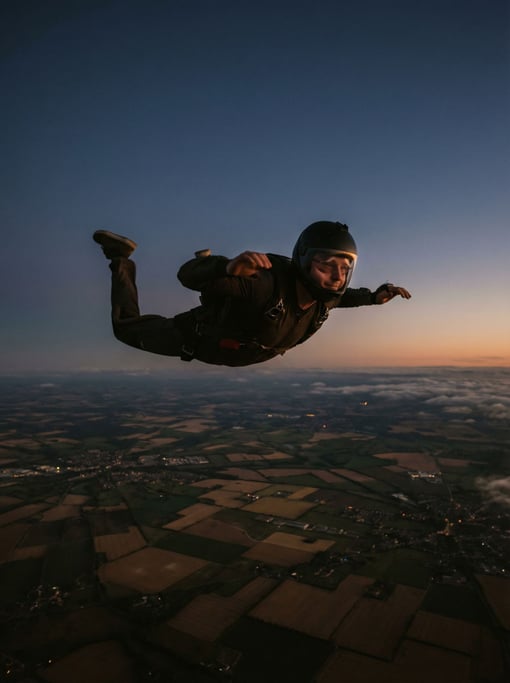 A skydiver in freefall, arms spread, earth a patchwork quilt far below, wind distorting cheeks