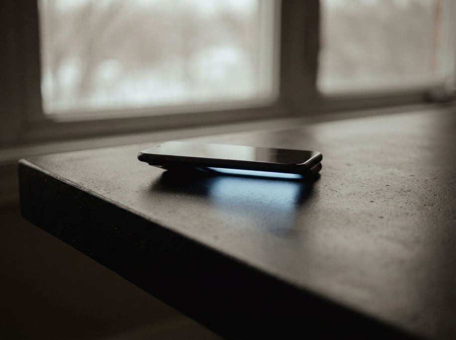 A smartphone face-down on a matte black table, screen glow casting a faint cool reflection upward (wn7lr7qf)