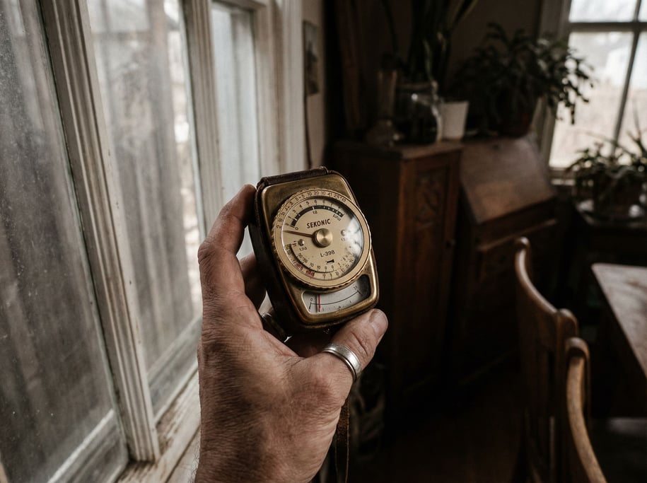 A light meter held up to a window, analog needle mid-swing, photographer's hand in frame