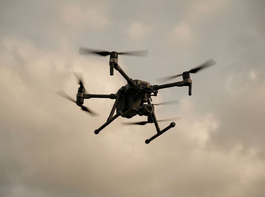 A drone mid-flight against an overcast sky, propellers blurred, body sharp, shot from below (sb)