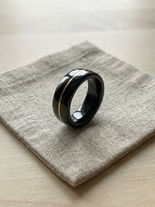 A concept smart ring in polished black ceramic with a hairline gold stripe inlay sits on raw linen