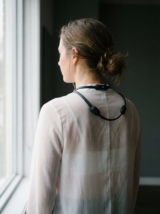 A woman wearing a concept posture-correcting wearable as a thin band across her upper back visible through a sheer white blouse