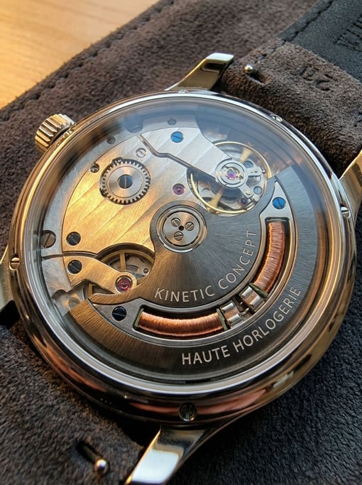 Gold-plated Geneva stripes catch warm light through a sapphire caseback — a concept kinetic energy harvesting watch lies face-down on dark grey suede