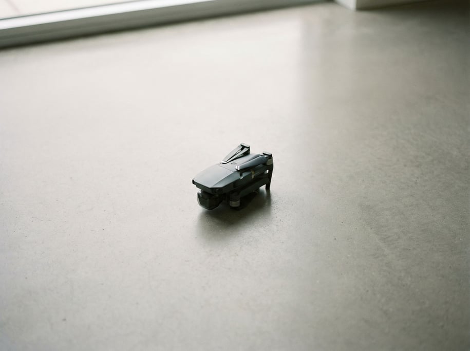 A drone folded on a clean concrete surface, propellers tucked, diffused light from above (vmpn)