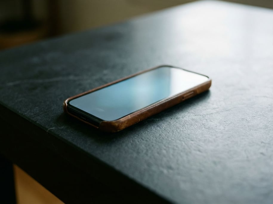 A smartphone face-down on a matte black table, screen glow casting a faint cool reflection upward