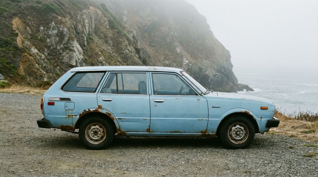 A weathered 1970s Japanese compact car in faded powder blue sits in a gravel lot beside a misty coas
