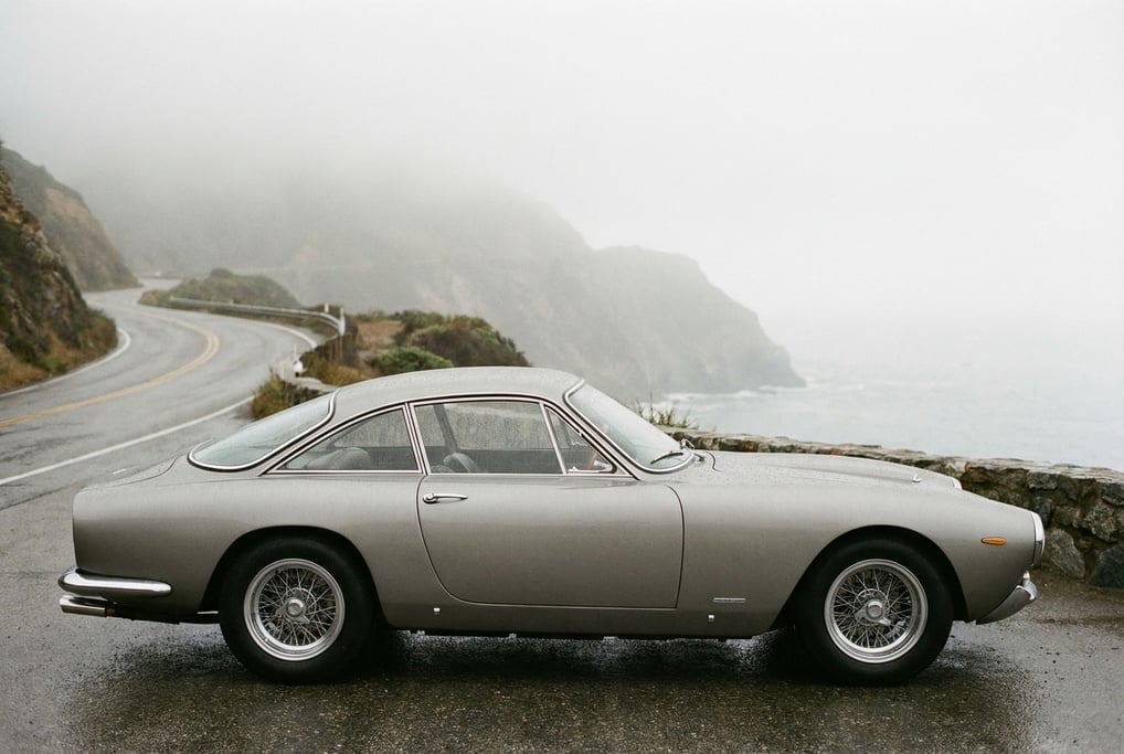 Side profile of a 1960s Italian grand tourer in dove grey on a misty coastal road