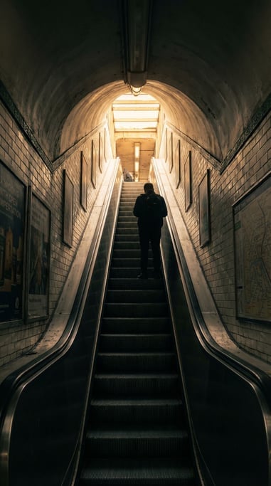 Subway escalator leading up to street level, warm daylight streaming down from the exit above