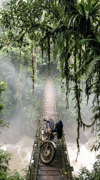 Motorcycle on a narrow bridge over a jungle river gorge, tropical vegetation hanging from above