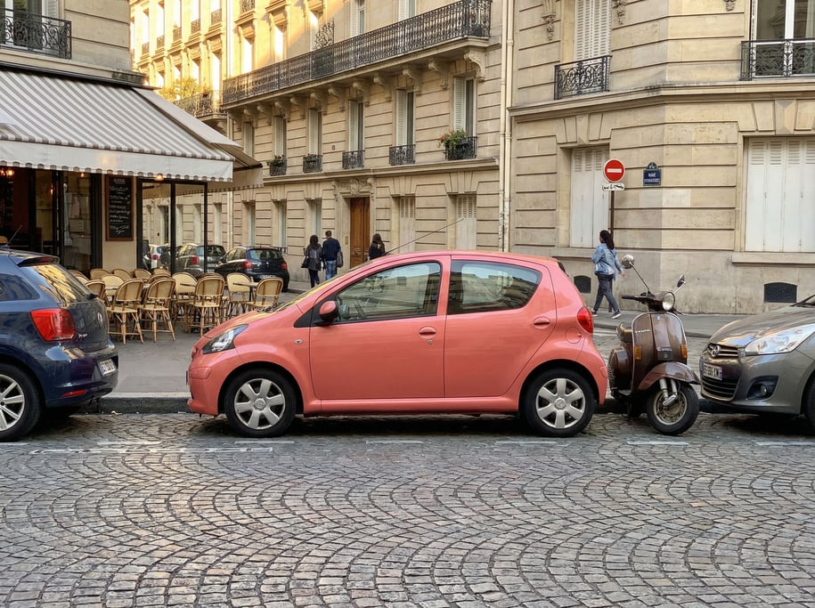 Small city car in coral pink wedged into a tight parallel parking spot on a Paris-style boulevard