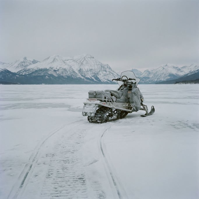 Snowmobile parked on a frozen lake, tracks in the snow behind it, mountains in the far background