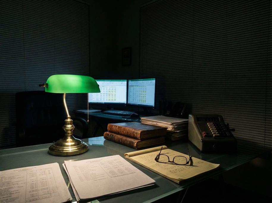 Green-tinted light from a brass banker's lamp carves a focused pool across a forensic accountant's g