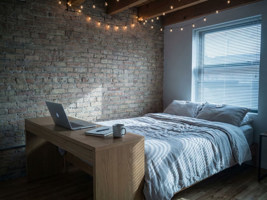 A loft bedroom workspace, laptop on a desk at the foot of an unmade bed, exposed brick wall (li)